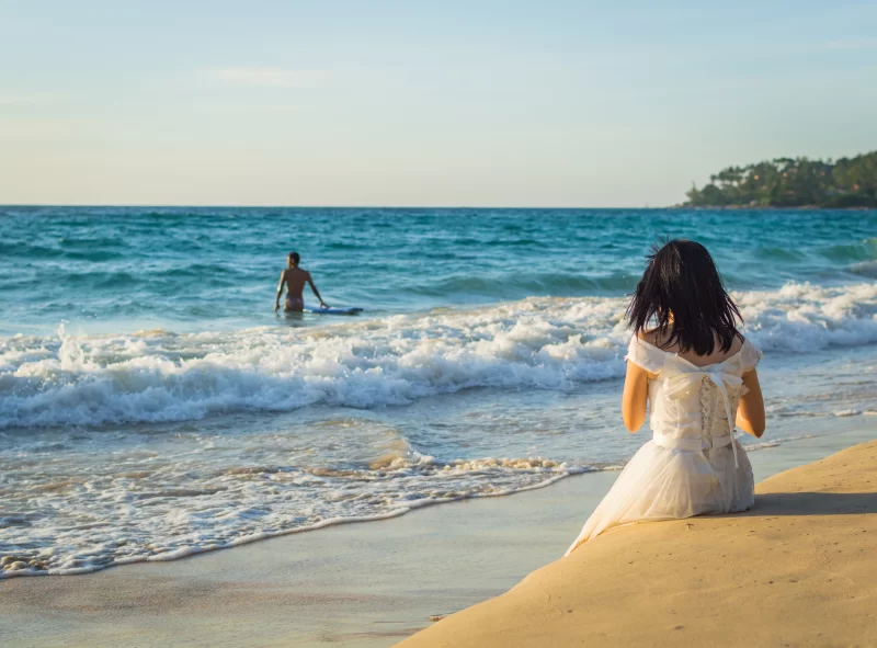 Девушка в белом платье на берегу моря, мужчина с доской в волнах