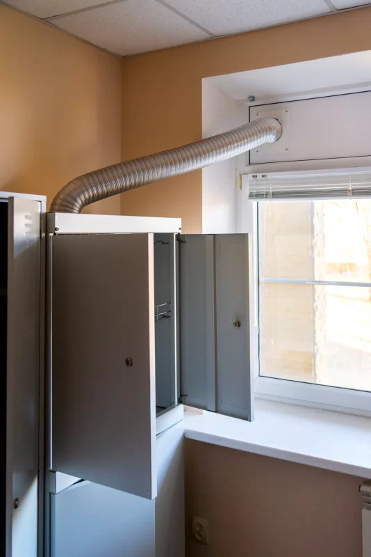 Вентиляционная труба, ведущая из шкафчика к окну, в помещении с бежевыми стенами