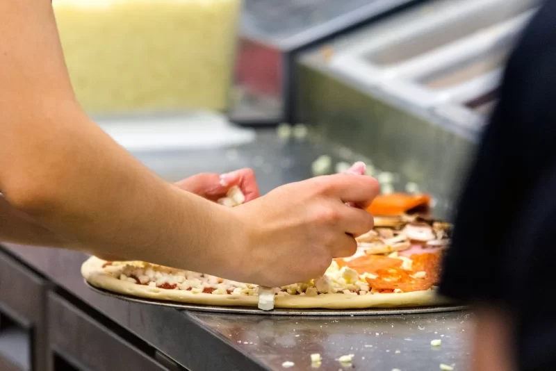 Руки повара готовят пиццу, добавляя сыр на основу с пепперони и грибами